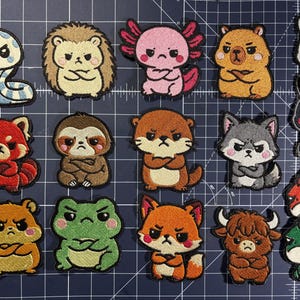 May include: A selection of embroidered patches showcasing a variety of grumpy cartoon animals. The collection includes a snake, hedgehog, axolotl, bear, rabbit, red panda, sloth, otter, wolf, hamster, frog, fox, bull, and dragon. Each patch has a unique design and colour scheme.