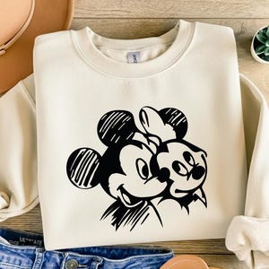 May include: Cream-colored sweatshirt featuring a black and white graphic of Mickey and Minnie Mouse. The design shows the iconic characters embracing. The sweatshirt has long sleeves and a crew neck, perfect for casual wear.