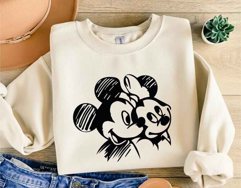 May include: Cream-colored sweatshirt featuring a black and white graphic of Mickey and Minnie Mouse. The design shows the iconic characters embracing. The sweatshirt has long sleeves and a crew neck, perfect for casual wear.