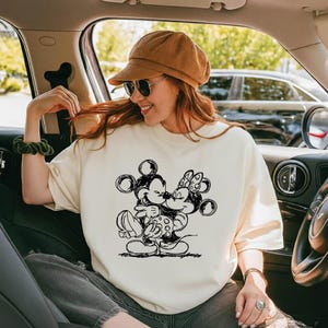 May include: Cream-colored sweatshirt featuring a black and white sketch of Mickey Mouse and Minnie Mouse embracing. The graphic is centered on the front of the shirt. The person in the image is wearing sunglasses and a brown corduroy cap.