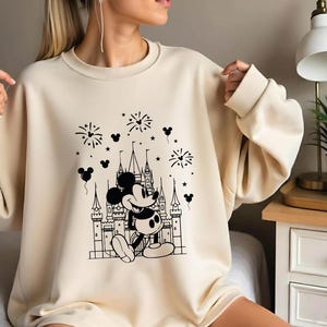 May include: Cream-colored sweatshirt featuring a black and white graphic of Mickey Mouse in front of a castle with fireworks and Mickey Mouse balloons. The sweatshirt has long sleeves and a crew neck.