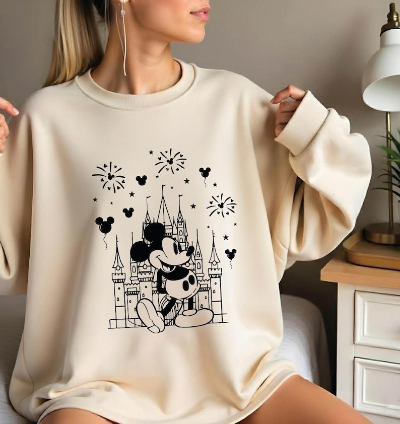 May include: Cream-colored sweatshirt featuring a black and white graphic of Mickey Mouse in front of a castle with fireworks and Mickey Mouse balloons. The sweatshirt has long sleeves and a crew neck.