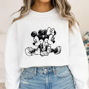 May include: White sweatshirt featuring a black and white sketch of Mickey Mouse and Minnie Mouse sitting together. The cartoon characters are in a relaxed pose, with Mickey holding a ball. The design is centered on the front of the shirt.