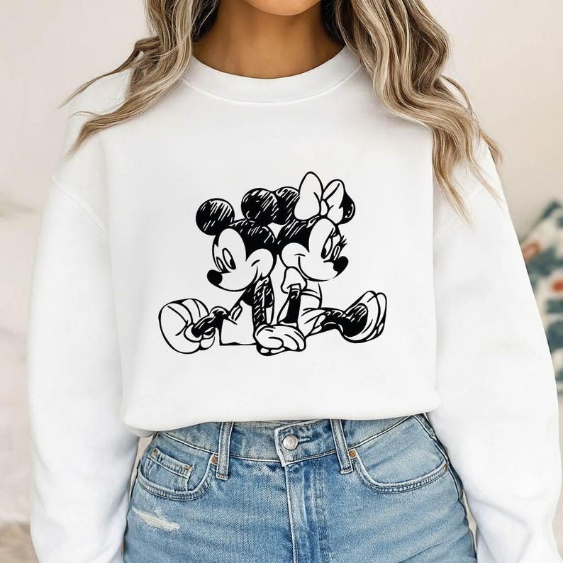 May include: White sweatshirt featuring a black and white sketch of Mickey Mouse and Minnie Mouse sitting together. The cartoon characters are in a relaxed pose, with Mickey holding a ball. The design is centered on the front of the shirt.