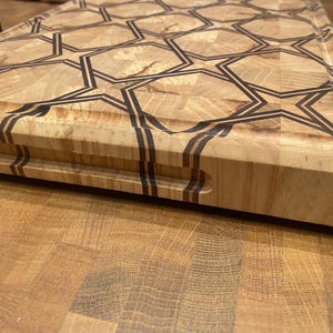 Puede incluir: Una tabla de cortar de madera con un patrón geométrico de estrellas. La tabla está hecha de madera clara con detalles en marrón oscuro. Tiene forma rectangular con un hueco para el asa en un lado. La tabla se utiliza probablemente para la preparación de alimentos.