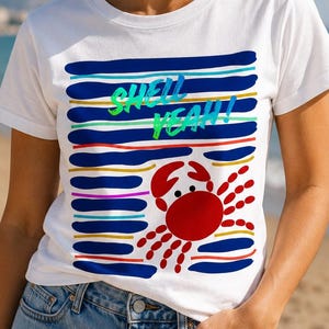 May include: A white short-sleeved t-shirt featuring a graphic print of a red crab and horizontal stripes in blue, yellow, and teal. The text "SHELL YEAH!" is printed in bright green and blue letters across the center of the striped pattern.