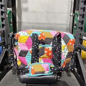 May include: A patchwork duffel bag in a variety of colours including pink, yellow, blue, and black geometric shapes. The bag has black straps with white stars and a blue accent. The bag is on a black surface.