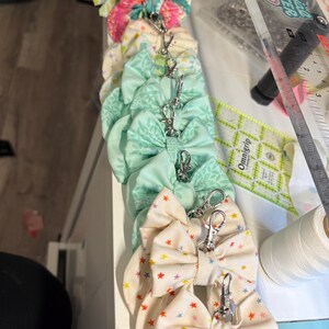 May include: A vertical row of fabric bows with silver metal lobster claw clasps. The bows feature various patterns including colorful floral prints, light green botanical designs, and a cream fabric with small multicolored stars.