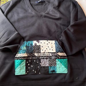 May include: A black V-neck sweatshirt featuring a large, quilted front pocket made of patchwork fabric. The pocket displays various patterns including black and white checkers, stars, and teal geometric shapes, with text reading sketchbook, mini starry, and star society.