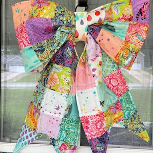 Pode incluir: Um grande laço de patchwork colorido, provavelmente para decoração. O laço apresenta uma variedade de padrões de tecido, incluindo desenhos florais, geométricos e abstratos em tons de rosa, verde, amarelo e roxo. É suspenso por um cabide de metal preto, adequado para uma porta ou janela.
