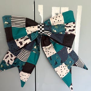 May include: A large fabric bow featuring a patchwork design of teal, black, and white squares. The patterns include black and white stripes, small black bats, stars, and black cats on a teal background. A black and white chequered knot centres the bow.