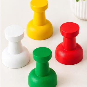 May include: A set of four wall hooks shaped like oversized push pins in solid white, yellow, red, and green colors. Each hook features a wide circular base that tapers into a narrow neck, topped with a flat, round head for hanging items.