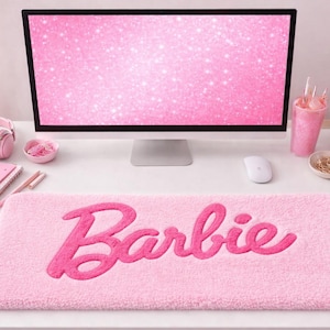 May include: A pink desk setup with a computer monitor displaying a pink glitter design, pink headphones, a notebook, and a mouse. A pink desk mat with the word "Barbie" in a script font is in the foreground.