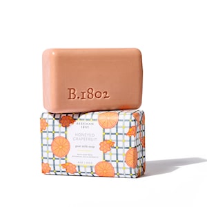 May include: A rectangular bar of peach-colored soap with the embossed text "B.1802" sits atop a patterned box. The box features a checkered design with orange grapefruit slices and the words "Honeyed Grapefruit goat milk soap".