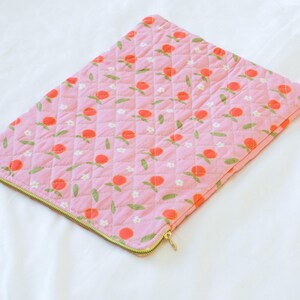 May include: Pink quilted laptop sleeve with a repeating pattern of orange flowers, green leaves, and white blossoms. The sleeve has a gold zipper closure and a quilted texture. The background is a white sheet.