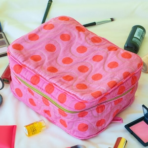 May include: A pink quilted makeup bag with a repeating pattern of large orange polka dots. The bag has a gold zipper and is surrounded by makeup products, including eyeshadow, blush, and brushes. The bag is rectangular and appears to be made of fabric.