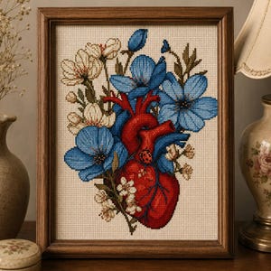 Pode incluir: Um bordado em ponto cruz emoldurado apresentando um coração anatômico vermelho vibrante cercado por cachos de flores azuis e brancas. A obra é bordada em um tecido bege claro e inserida em uma moldura de madeira marrom escura.