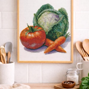 Puede incluir: Obra de arte de punto de cruz de un tomate, una col y zanahorias en un marco de madera. Las verduras están representadas en colores vibrantes, con el tomate de un rojo intenso y la col una mezcla de verde y morado. Las zanahorias son naranjas. La obra se exhibe en una cocina.
