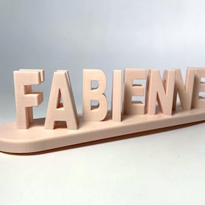 Peut inclure: Une enseigne tridimensionnelle rose clair affichant le nom FABIENNE en lettres capitales. Les lettres sont fixées sur une base lisse de forme ovale de la même couleur, formant un objet décoratif posé avec une finition mate.