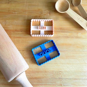 Peut inclure: Deux emporte-pièces rectangulaires aux bords festonnés, l'un rose clair avec le mot aim et l'autre bleu avec le mot pond. Un rouleau à pâtisserie en bois et deux cuillères à mesurer en bois, dont une marquée 15 ml, sont placés à proximité.