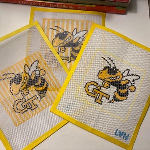 May include: Three needlepoint canvases with a yellow and black bee design, featuring the letters "GT". The canvases have a white background with yellow borders. The bee is wearing sunglasses. The initials "LVN" are in the bottom right corner.