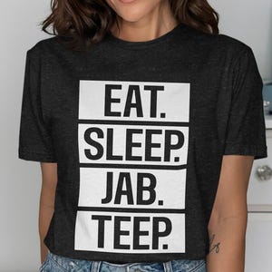Puede incluir: Una camiseta de manga corta en color gris carbón oscuro con cuatro bloques rectangulares blancos apilados con texto negro. El texto dice EAT. SLEEP. JAB. TEEP. en una fuente sans-serif en negrita. La camiseta tiene cuello redondo.