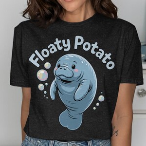 May include: A dark charcoal gray short-sleeve t-shirt featuring a light blue cartoon manatee illustration with pink cheeks. The text "Floaty Potato" is printed in light blue above the manatee, which is surrounded by several small bubbles.