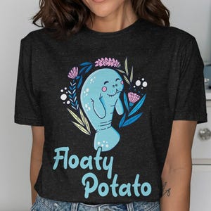 Könnte beinhalten: Ein dunkel anthrazitfarbenes T-Shirt mit einer hellblauen Seekuh-Illustration, umgeben von rosa und blauen floralen Elementen. Unter der Grafik ist der Text "Floaty Potato" in einer hellblauen, abgerundeten serifenlosen Schrift aufgedruckt.