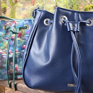 May include: Two drawstring bucket bags. One is navy blue leather, the other is green with a floral print reminiscent of a Monet painting. Both have silver-toned hardware and drawstring closures. The bags are displayed outdoors.