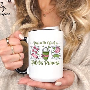 May include: White ceramic mug with a black handle and rim, featuring the text "Day in the Life of a Pilates Princess." The design includes illustrations of dumbbells, a yoga mat, a drink, socks, and a sleep mask. The mug has the "mugsursprise" logo.