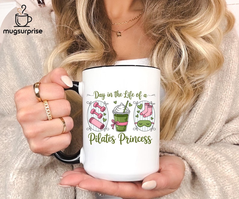 May include: White ceramic mug with a black handle and rim, featuring the text "Day in the Life of a Pilates Princess." The design includes illustrations of dumbbells, a yoga mat, a drink, socks, and a sleep mask. The mug has the "mugsursprise" logo.