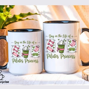 May include: Two white ceramic mugs with black rims and handles. Each mug has the text "Day in the Life of a Pilates Princess" with illustrations of dumbbells, a drink, socks, and a sleep mask. The mugs are on a light-coloured surface.