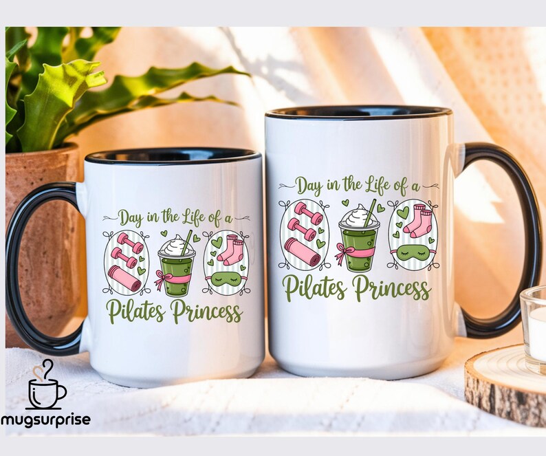May include: Two white ceramic mugs with black rims and handles. Each mug has the text "Day in the Life of a Pilates Princess" with illustrations of dumbbells, a drink, socks, and a sleep mask. The mugs are on a light-coloured surface.