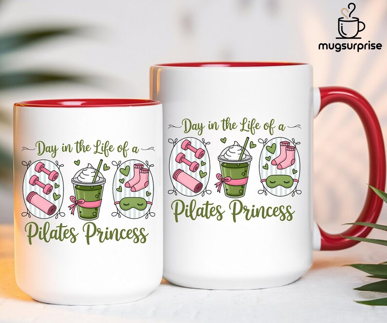 May include: Two white ceramic mugs with red interiors and handles. The mugs feature the text "Day in the Life of a Pilates Princess" with illustrations of dumbbells, a drink, socks, and a sleep mask. The mugs are for beverages.