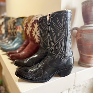 May include: A row of cowboy boots in various colors and designs. The front pair is black leather with white stitching. Other boots include blue, red, and brown, with floral and geometric patterns. The boots are displayed on a white shelf.