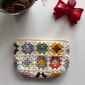 May include: A crochet pouch with a floral design in various colours. The pouch is made with cream-coloured yarn and has a zip closure. The floral squares are in shades of pink, blue, green, yellow, and purple. A red bow-shaped candle holder is also visible.