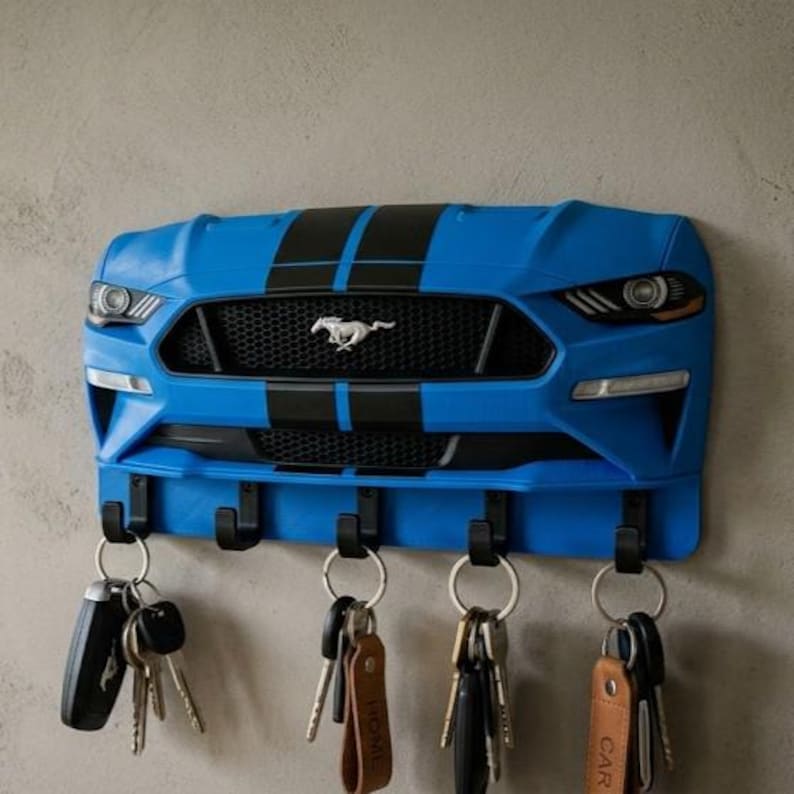 May include: A blue wall-mounted key holder shaped like the front end of a Ford Mustang car, featuring a black grille, a silver horse emblem, and two black racing stripes. It includes five black hooks holding various metal key rings and key fobs.