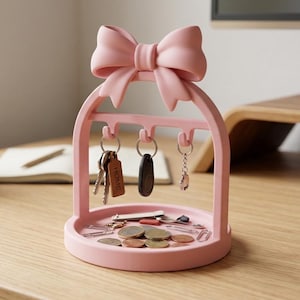 May include: A light pink arched key holder featuring a large decorative bow at the top and three hooks for hanging keys. The base is a circular tray containing various metal paper clips, coins, and a small rectangular tag labelled HOME.