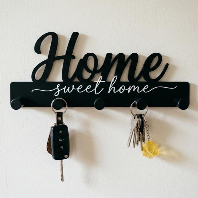 May include: A black wall-mounted key holder featuring the word Home in a large script font above the text sweet home in a smaller script font. The rectangular base has five cylindrical pegs, with a car key fob and a set of keys attached.