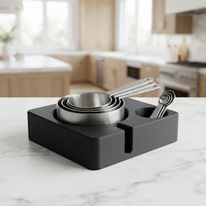 May include: A set of stainless steel measuring cups and spoons nested within a matte black rectangular storage base. The base features a dedicated circular cutout for the cups and a smaller circular slot for the spoons, with a vertical notch.