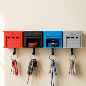 May include: Four wall-mounted garage-style key holders in red, black, blue, and grey. Each unit features a miniature car inside, a garage door design, and the text "GARAGE KEY HOLDER" embossed on top. A black hook hangs below each, holding various metal keys.