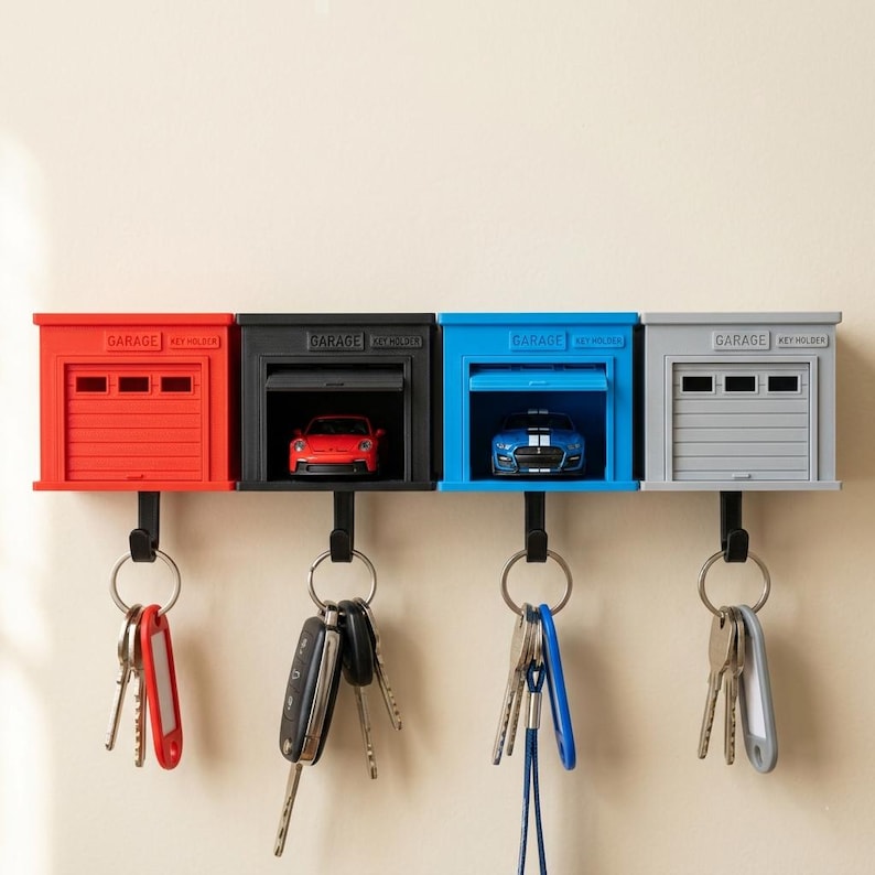 May include: Four wall-mounted garage-style key holders in red, black, blue, and grey. Each unit features a miniature car inside, a garage door design, and the text "GARAGE KEY HOLDER" embossed on top. A black hook hangs below each, holding various metal keys.