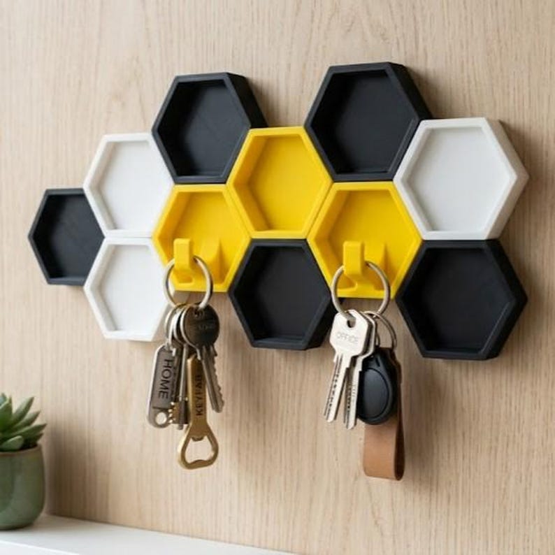 May include: A wall-mounted key holder featuring a cluster of interlocking hexagonal modules in black, white, and yellow. Two yellow modules include integrated hooks, each holding a set of metal keys with various tags and a bottle opener.