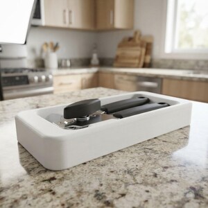 May include: A black manual can opener with a rotating handle and a metal cutting mechanism, resting inside a rectangular white storage tray. The tool features a matte black finish on the handles and a metallic silver gear for opening cans.