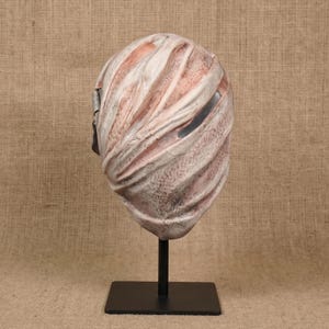 Op de afbeelding: Een masker voor het hele hoofd met een textuur van windsel in tinten gebroken wit, beige en roodbruin. Het oppervlak heeft diepe, diagonale groeven en plooien die op verband lijken, met een enkele smalle, donkere horizontale oogopening.