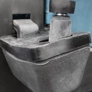 Könnte beinhalten: Nahaufnahme einer schwarzen, industriell aussehenden Komponente mit einem zylindrischen Pfosten und einer flachen, rechteckigen Basis. Ein kleineres, kreisförmiges Teil sitzt auf dem Pfosten, befestigt durch einen silbernen Verschluss. Die Komponente scheint aus einem haltbaren, möglicherweise gummierten Material zu bestehen.