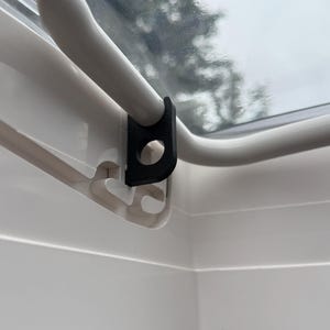 Op de afbeelding: Close-up van een zwarte kunststof raamvergrendeling bevestigd aan een wit raamkozijn. De vergrendeling is ontworpen om een raam of ventilatieopening te beveiligen, met een gebogen handvat en een cirkelvormige opening. De achtergrond toont een wazig zicht op bomen en een bewolkte lucht.