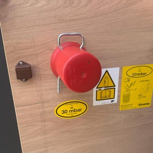 Può includere: Un regolatore di gas rosso con maniglia in metallo è montato su una superficie in legno marrone chiaro. Sono visibili due adesivi gialli con testo e simboli neri, insieme a un piccolo fermo rettangolare marrone.