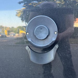 Könnte beinhalten: Eine dunkelgraue, kreisförmige Truma-Lüftungsabdeckung mit dem eingeprägten Wort HOT auf der mittleren Kappe. Unter der runden Lüftung befindet sich eine dunkelgraue, gebogene, 3D-gedruckte Kunststoffhalterung auf einer dunklen, reflektierenden Oberfläche.