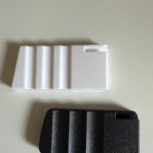 Könnte beinhalten: Zwei Kunststoffkomponenten, eine weiße und eine schwarze, werden gezeigt. Das weiße Teil hat eine geriffelte Oberfläche und einen rechteckigen Ausschnitt. Das schwarze Teil hat ebenfalls eine geriffelte Oberfläche und eine kleine Schraube. Beide Teile sind rechteckig.